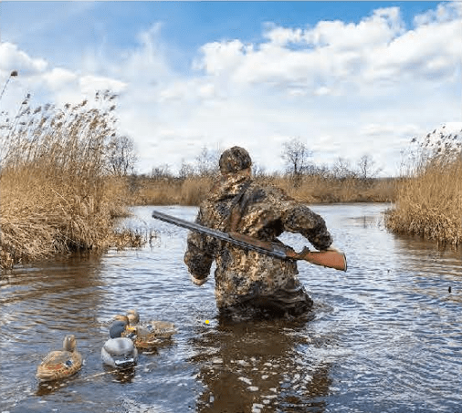 A camouflaged hunter wades through deep water, duck decoys trailing behind