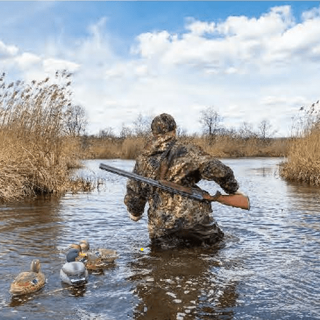 A camouflaged hunter wades through deep water, duck decoys trailing behind